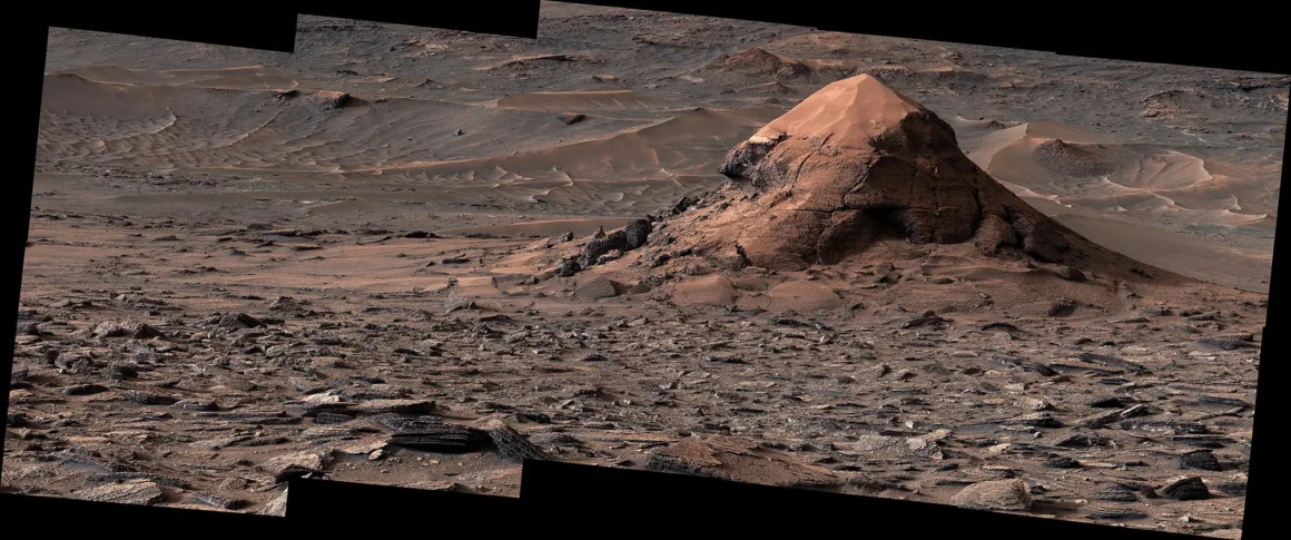 Kayalık ve çorak Mars arazisinde, kızıl tozla kaplı belirgin koni biçimli bir tepe ve çevresine yayılmış kaya parçaları görülüyor.
