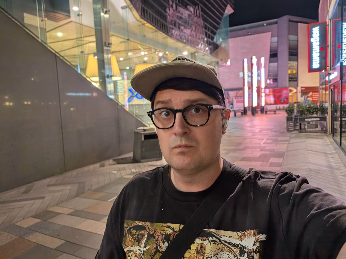 Google Pixel 11 Pro Fold İncelemesi: Şık Ama Yerinde Sayıyor 39 Aydınlatılmış tabelaların bulunduğu yaya alışveriş caddesinde gece selfie’si çeken, gözlüklü ve beyzbol şapkalı adam.