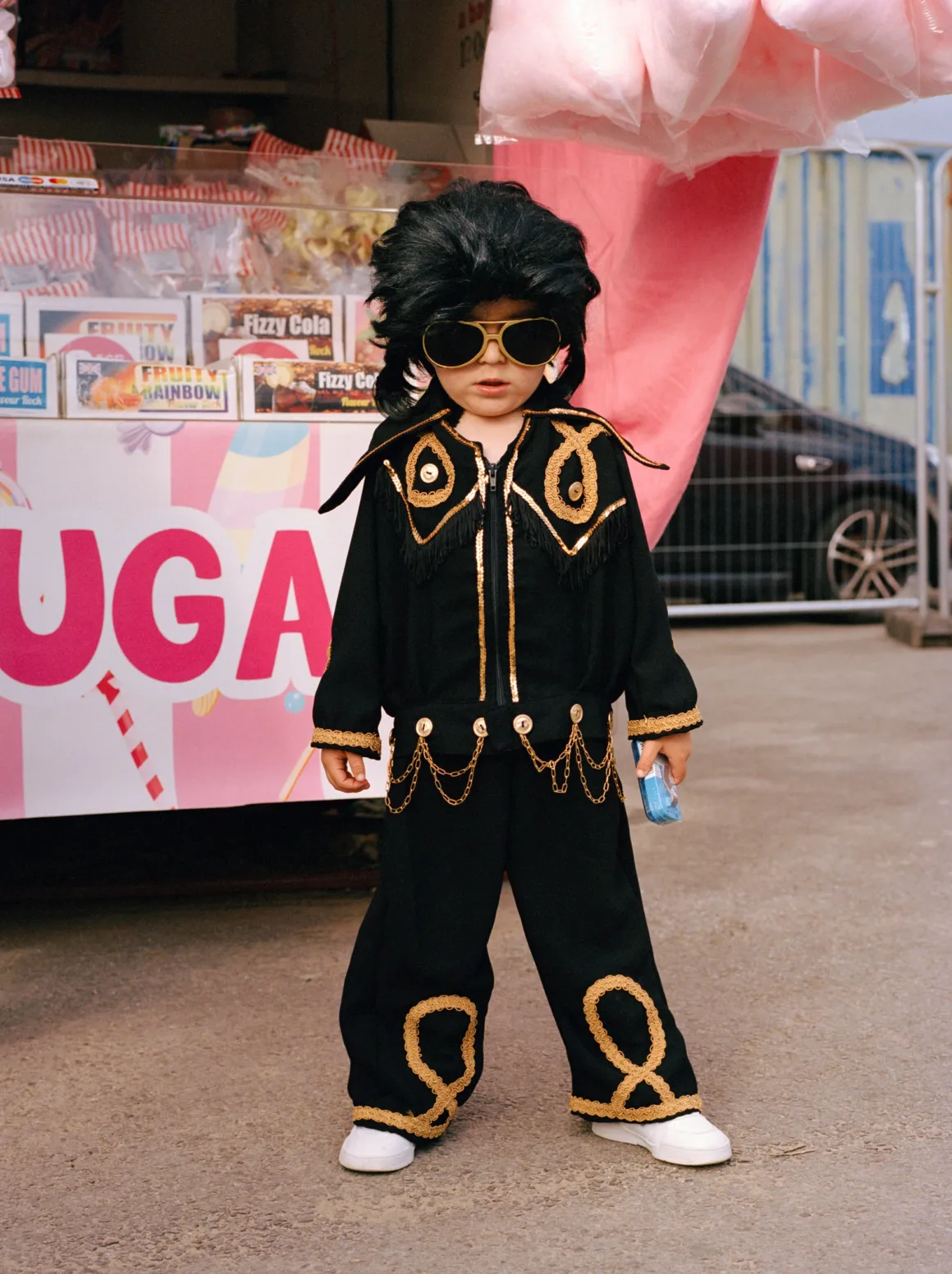 Winston Taylor Photo Portrait Prize 2026 8 A young child dressed in an Elvis Presley costume, including a black jumpsuit with gold trim, a black wig, and sunglasses, stands in front of a candy stand.