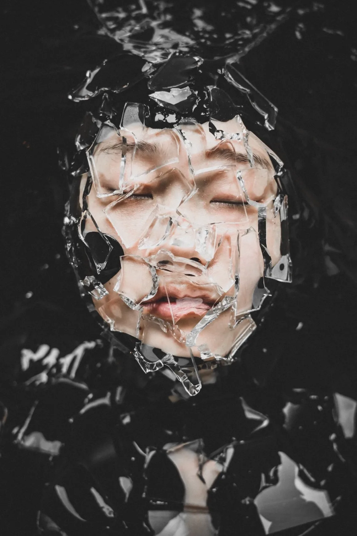 2026 European Photography Awards: Dünyayı Görmenin Farklı Yolları 23 Bir kişinin yüzü kırık cam parçaları tarafından kısmen örtülmüş; gözleri karanlık ve sıvıyı andıran bir arka plan önünde kapalı.