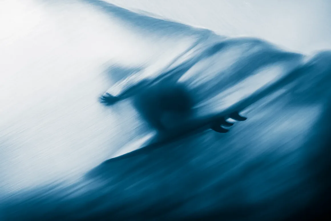 Okyanusun Görünmeyen Yüzü: Ocean Photographer of the Year 2026 Finalistleri 28 Mavi tonlardaki bulanık ve soyut sahnede bir sörfçü dalganın üzerinde görülüyor; akışkan hareket etkisi dinamik hareketi vurguluyor.