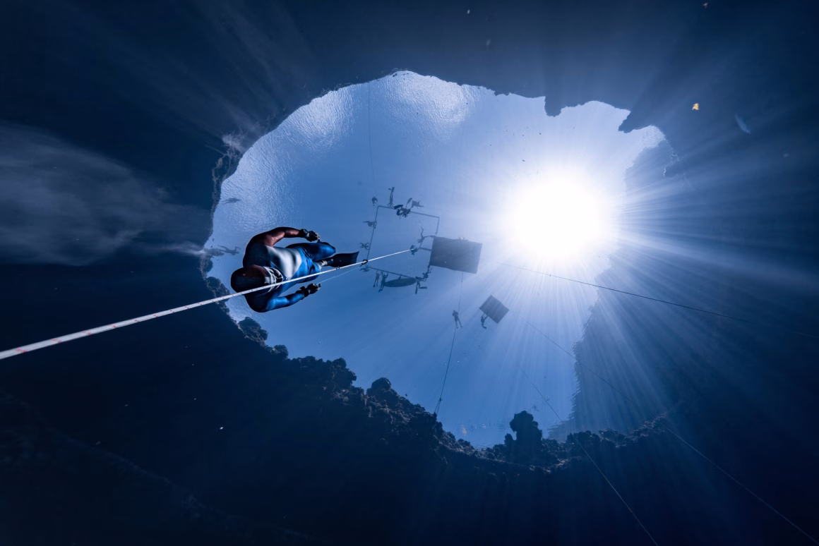 Okyanusun Görünmeyen Yüzü: Ocean Photographer of the Year 2026 Finalistleri 29 Serbest dalgıç, su yüzeyine doğru uzanan bir ip boyunca yükseliyor; yukarıdaki dairesel açıklıktan güneş ışığı süzülürken berrak mavi suda sualtı ekipmanları yüzüyor.