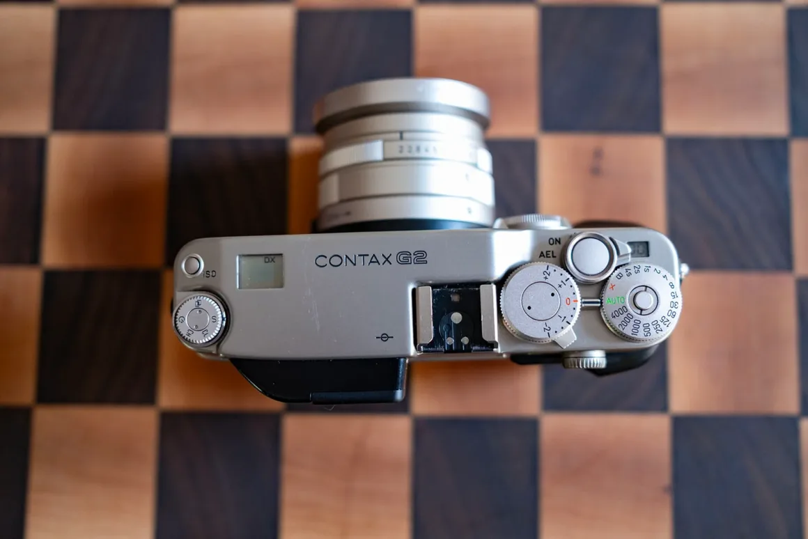 Contax G2 Efsanesi Boşuna Değil: Bu Kamera Harika 16 Objektifi takılı, gümüş renkli bir Contax G2 fotoğraf makinesi ahşap dama desenli bir yüzey üzerinde üstten görülüyor.