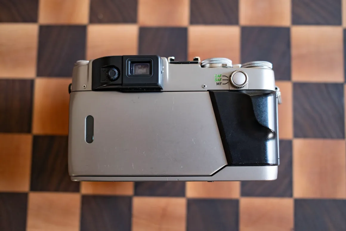 Contax G2 Efsanesi Boşuna Değil: Bu Kamera Harika 17 Ahşap bir satranç tahtası üzerinde duran gümüş ve siyah renkli bir analog fotoğraf makinesi arkadan görülüyor; vizörü, netleme modu seçicisi ve siyah tutma yeri dikkat çekiyor.