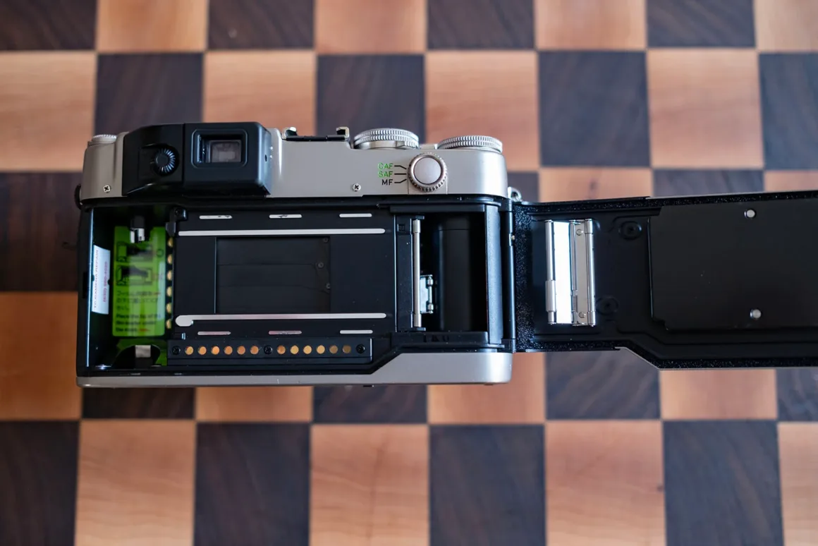 Contax G2 Efsanesi Boşuna Değil: Bu Kamera Harika 20 Ahşap dama desenli bir yüzey üzerinde duran açık durumdaki 35 mm analog fotoğraf makinesinin film haznesi, deklanşör perdeleri ve sarma makarası görülüyor.