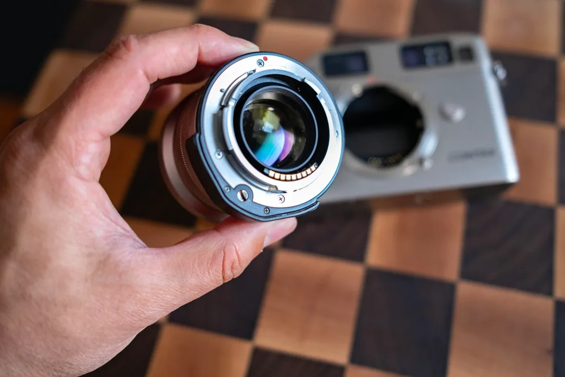 Contax G2 Efsanesi Boşuna Değil: Bu Kamera Harika 25 Bir el, dama desenli ahşap yüzey üzerinde duran gümüş renkli fotoğraf makinesi gövdesinin önünde, elektronik bağlantı noktaları görülen bir objektif tutuyor.