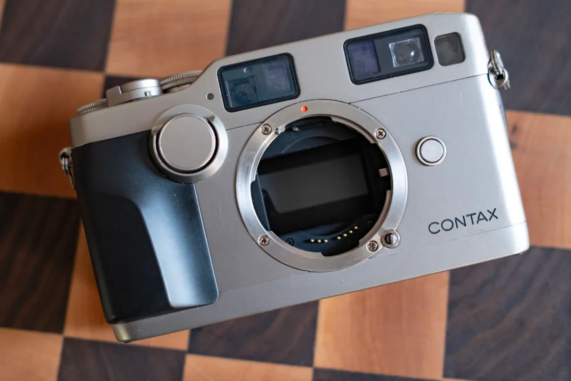 Contax G2 Efsanesi Boşuna Değil: Bu Kamera Harika 37 Objektif yuvası, vizör pencereleri ve siyah tutma yeri bulunan gümüş renkli bir Contax fotoğraf makinesi gövdesi, ahşap dama desenli bir yüzey üzerinde duruyor.