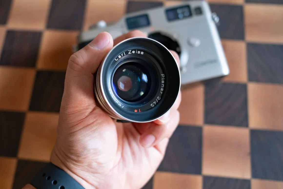 Contax G2 Efsanesi Boşuna Değil: Bu Kamera Harika 24 Bir el, dama desenli yüzey üzerinde duran gümüş renkli Contax fotoğraf makinesi gövdesinin önünde Carl Zeiss Planar 2/45 objektifi tutuyor.