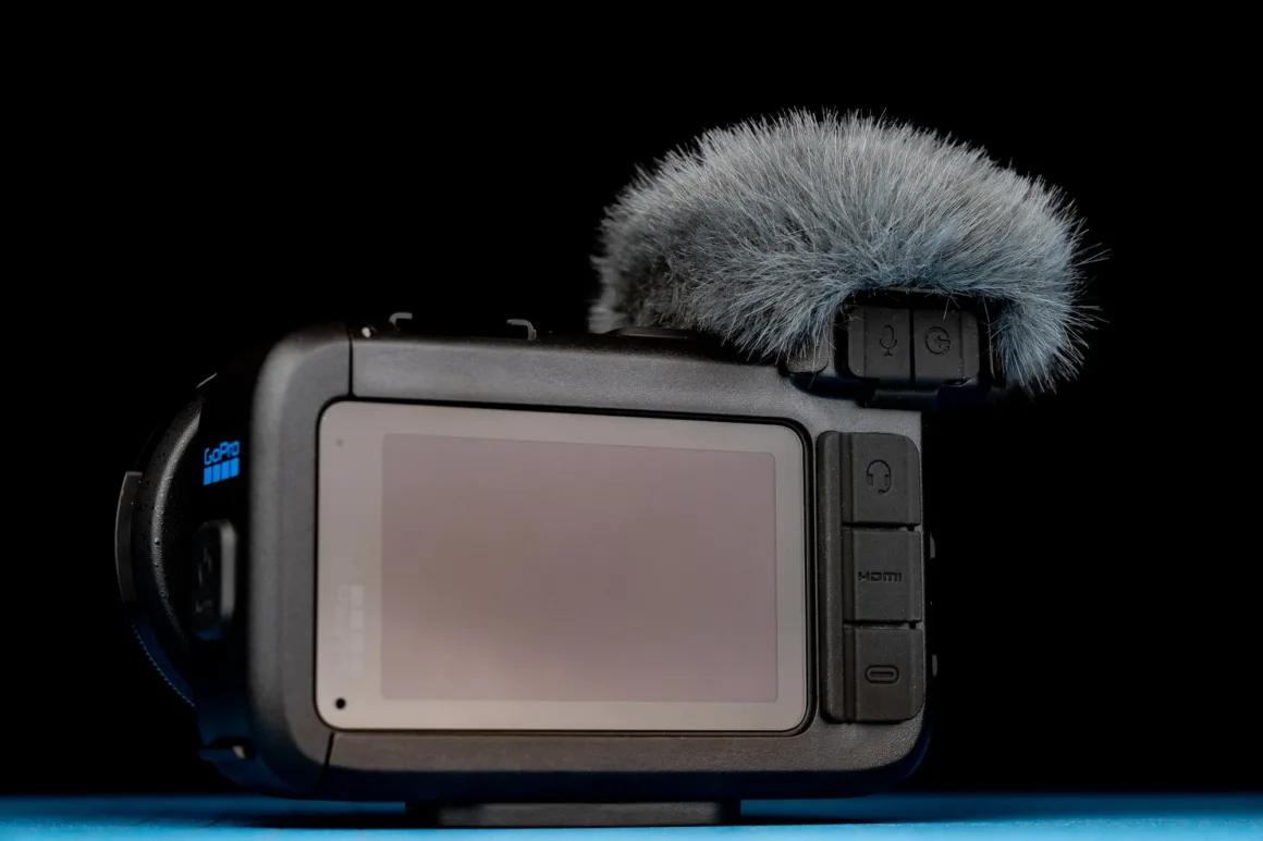 GoPro Mission 1 Pro ILS İncelemesi: Yenilikçi Ama Kime Göre? 25 Üstünde gri mikrofon rüzgârlığı bulunan siyah GoPro kamera, mavi yüzey üzerinde siyah fon önünde duruyor.