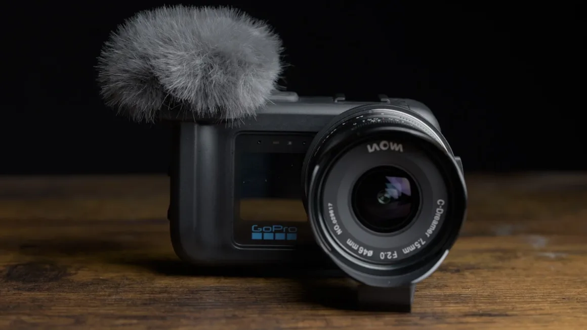 GoPro Mission 1 Pro ILS İncelemesi: Yenilikçi Ama Kime Göre? 21 Laowa C-Dreamer 7,5 mm objektif ve tüylü mikrofon rüzgârlığı takılı GoPro kamera, siyah fon önündeki ahşap yüzeyde duruyor.