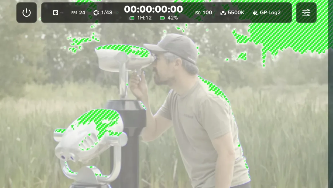 GoPro Mission 1 Pro ILS İncelemesi: Yenilikçi Ama Kime Göre? 28 Çimenlik alandaki sabit dürbünden bakan bir kişi; görüntünün parlak bölgelerinde yeşil zebra pozlama uyarıları görülüyor.