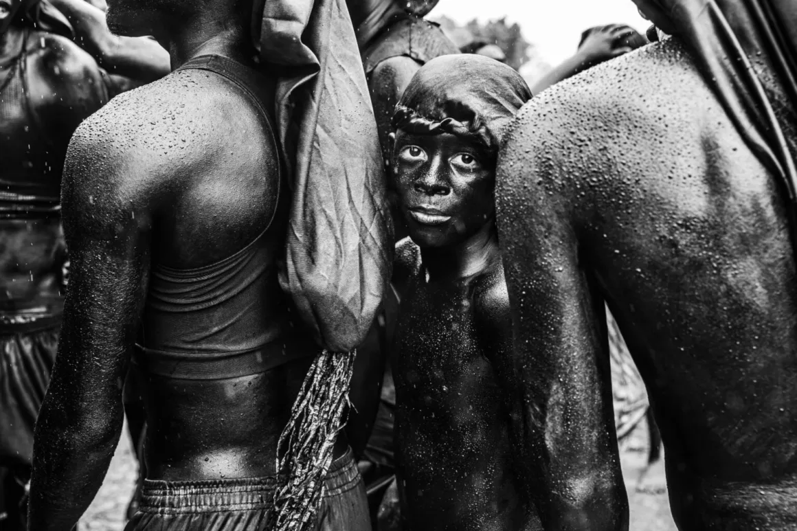 Geçip Giden Anlar, Sıra Dışı Bakışlar: Pure Street Photography Awards 2026 Kazananları
Yüzü koyu renge boyanmış ve başında örtü bulunan küçük bir çocuk, yağmur altında ıslak ve parıldayan tenleriyle iki yetişkinin arasında duruyor.