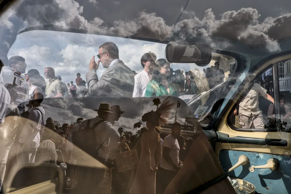 Çift pozlama tekniğiyle çekilmiş fotoğrafta, açık havadaki bir insan kalabalığı; pencere çerçevesi ve kapı kolu da görülen klasik bir otomobilin iç mekânıyla üst üste bindirilmiş.