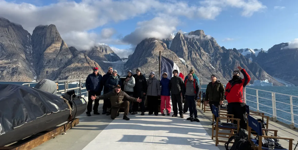 Scoresby Sund’da Işıklar Söndüğünde: Arktik’te Tam Güneş Tutulması 25 Tutulma gözlükleri takan bir grup insan, arkalarındaki geniş, dağlık ve karla kaplı manzara önünde bir teknenin güvertesinde duruyor.