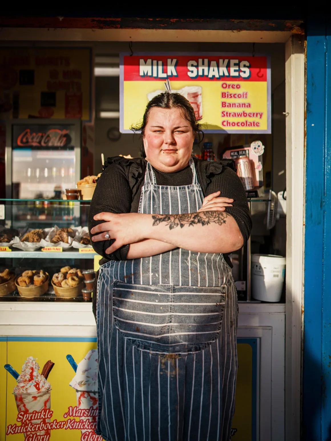 Winston Taylor Photo Portrait Prize 2026 13 A person with a floral forearm tattoo and a striped apron stands with arms crossed in front of a food stall advertising milkshakes and donuts.