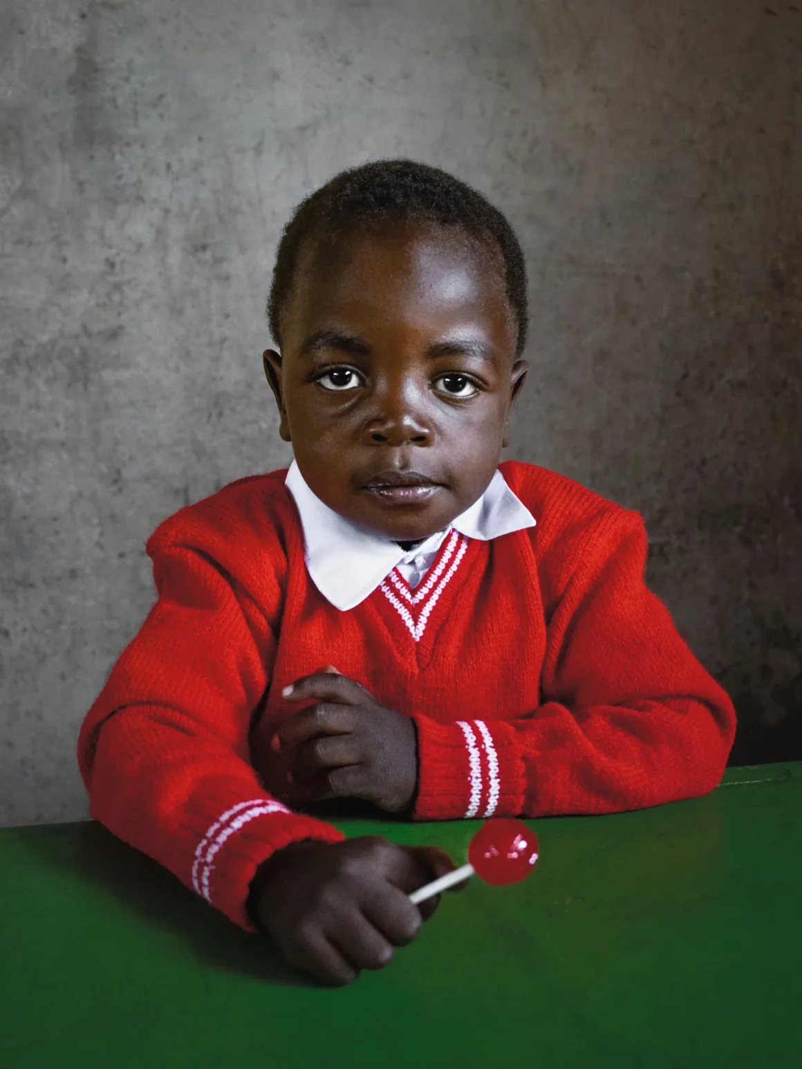 Winston Taylor Photo Portrait Prize 2026 14 A young child with dark skin wears a red sweater with a white collar and holds a red lollipop while sitting at a green desk.