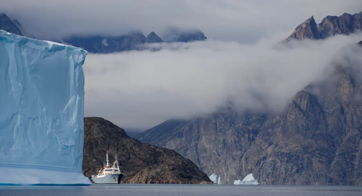 Scoresby Sund’da Işıklar Söndüğünde: Arktik’te Tam Güneş Tutulması 19 Beyaz bir keşif gemisi, sivri ve bulutlarla kaplı dağların önünde, devasa mavi bir buzdağının yanından sakin sularda ilerliyor.