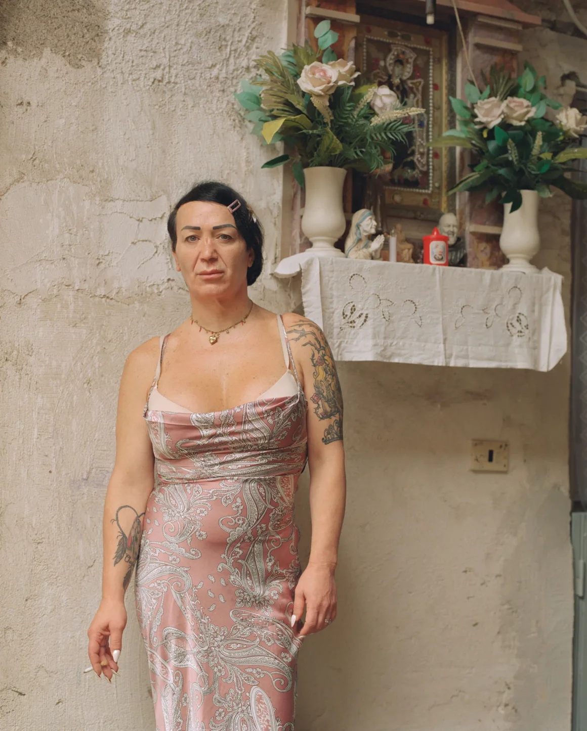Winston Taylor Photo Portrait Prize 2026 15 A person with dark hair and tattoos stands in a pink paisley dress, holding a cigarette, in front of a wall-mounted religious shrine with flowers.
