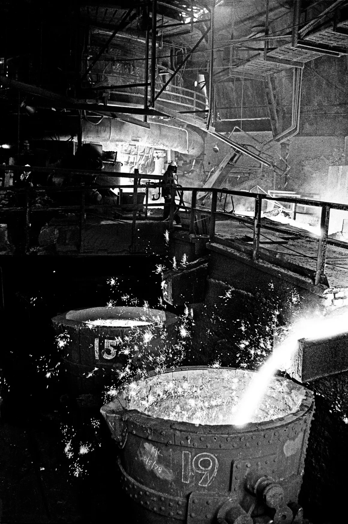 Erimiş metal büyük bir endüstriyel hazneye dökülürken etrafa kıvılcımlar saçılıyor. Ağır makineler ve borularla dolu karanlık ve sert fabrika ortamında, arka planda korkulukların yakınında bir işçi duruyor.