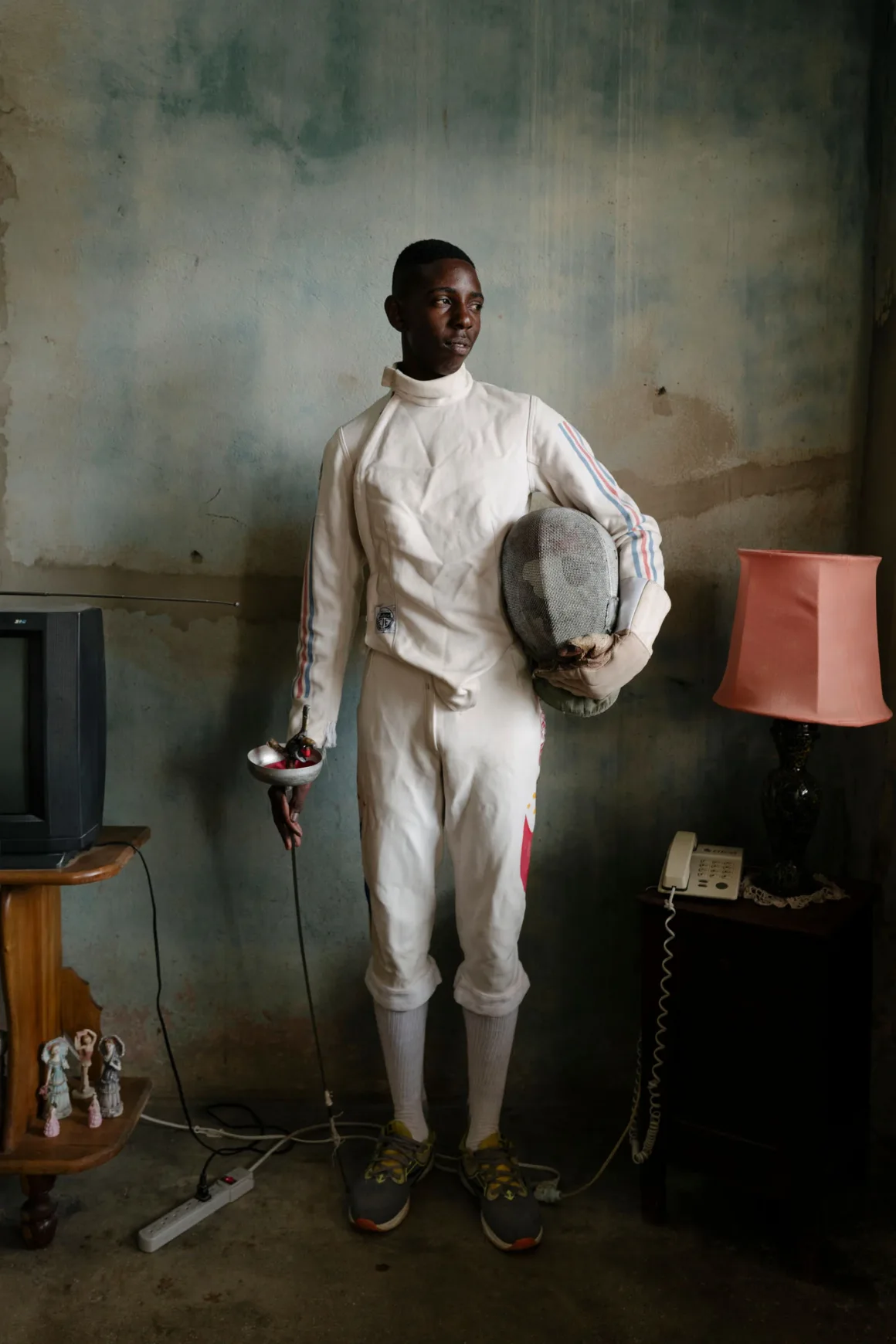 Winston Taylor Photo Portrait Prize 2026 4 Beyaz üniformalı genç bir eskrimci, televizyon, lamba ve eski bir telefonun bulunduğu odada maskesini ve flöresini tutarak ayakta duruyor.