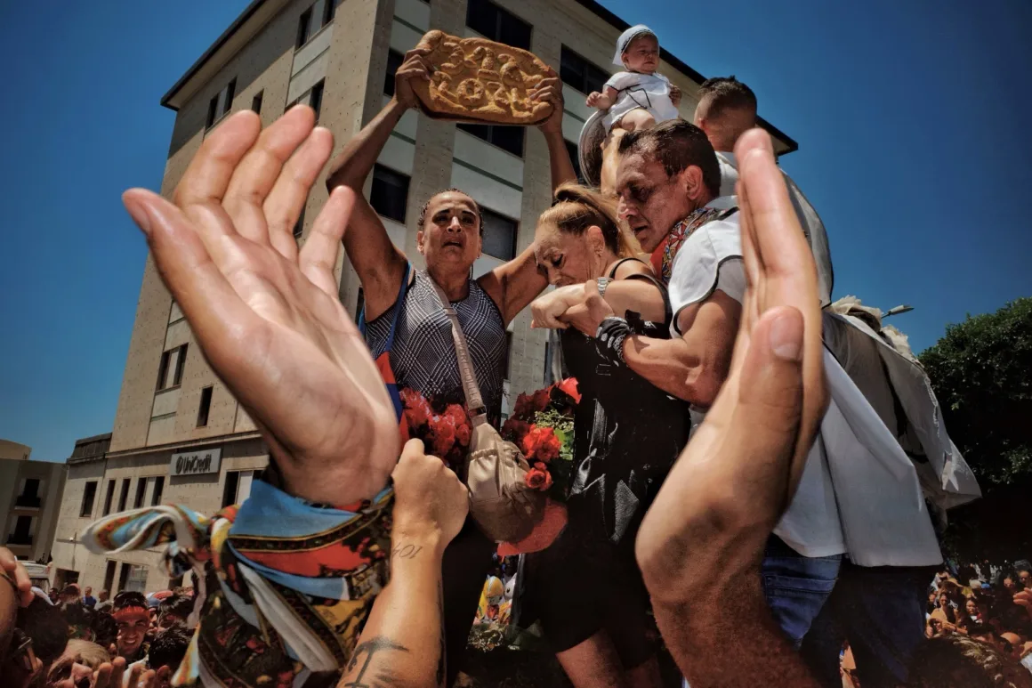 Alçak açıdan çekilen fotoğrafta bir insan kalabalığı görülüyor; ön planda havaya kalkmış eller, başının üzerinde büyük ve süslü bir ekmek taşıyan kadını çevreliyor.