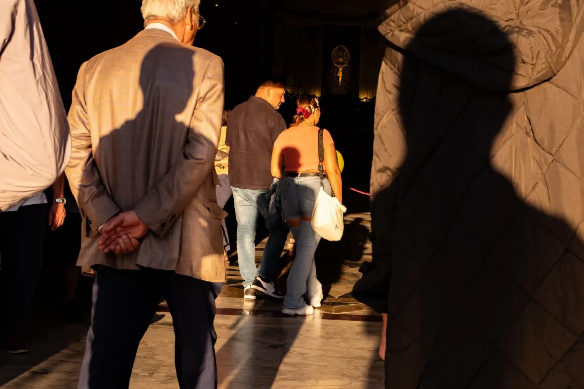 İnsanlar, duvarda uzakta aydınlatılmış bir haça doğru, güneş ışığı ve gölgelerin biçimlendirdiği kapalı bir mekânda yürüyor.