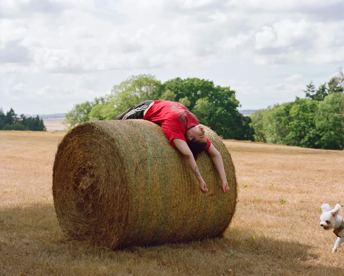 Winston Taylor Photo Portrait Prize 2026 16 A person in a red t-shirt and black athletic shorts lies arched backward over a large round hay bale in a field, while a small white dog walks nearby.