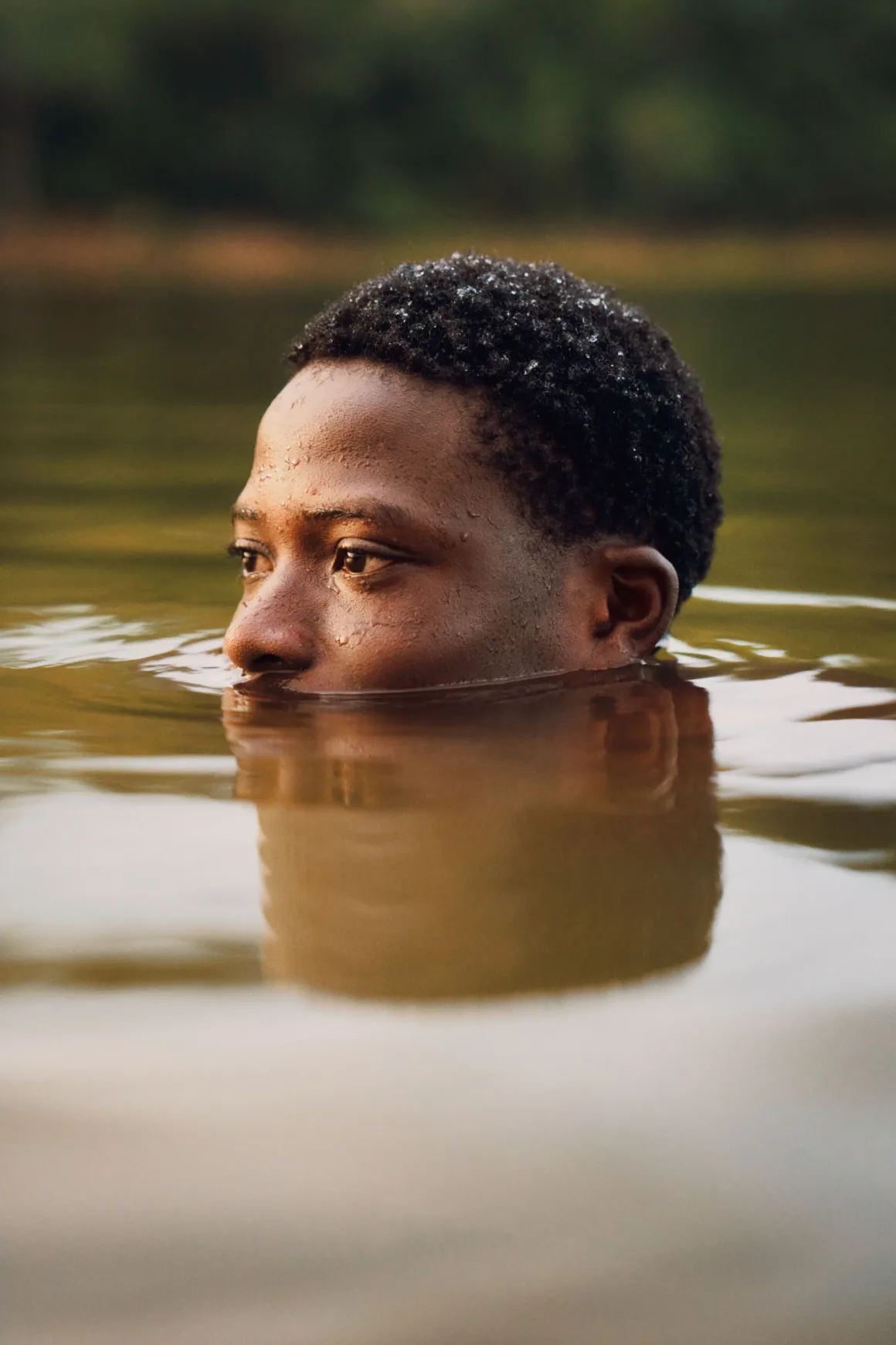 Winston Taylor Photo Portrait Prize 2026 18 A person with short, dark, textured hair is partially submerged in calm, murky water, looking off to the side.