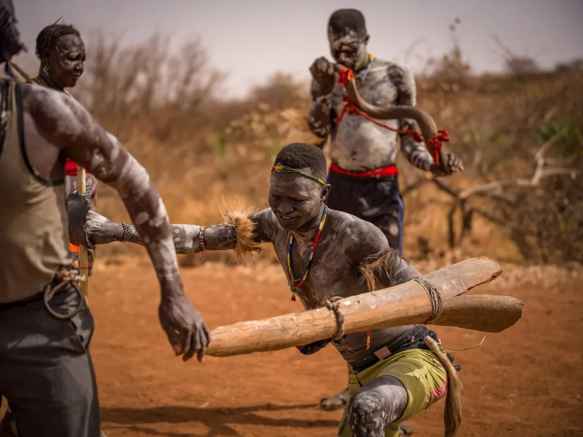 2026 European Photography Awards: Dünyayı Görmenin Farklı Yolları 27 Vücut boyaları ve geleneksel süslemeler taşıyan üç erkek, kuru ve açık bir alanda törensel bir etkinliğe katılıyor.
