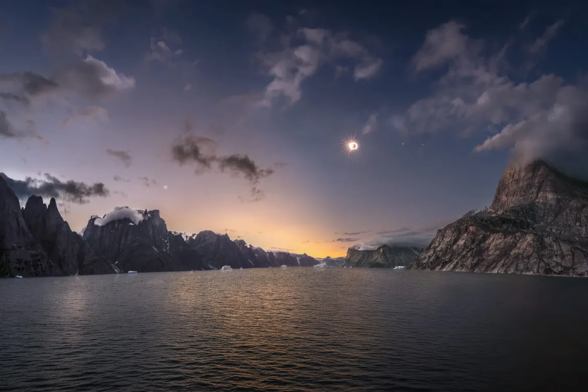 A total solar eclipse hangs in the sky above a calm body of water surrounded by jagged, cloud-capped mountains.