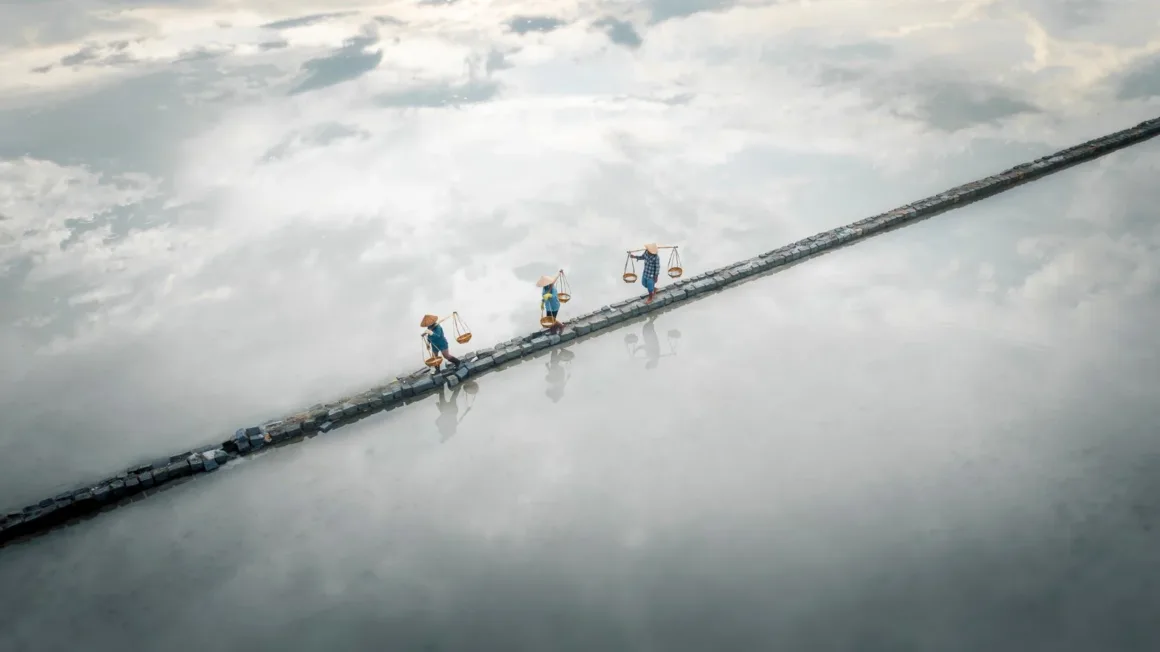 2026 European Photography Awards: Dünyayı Görmenin Farklı Yolları 20 Konik şapkalar takan ve omuz sırıklarında sepetler taşıyan üç kişi, su üzerindeki dar bir taş yolda tek sıra halinde yürüyor.