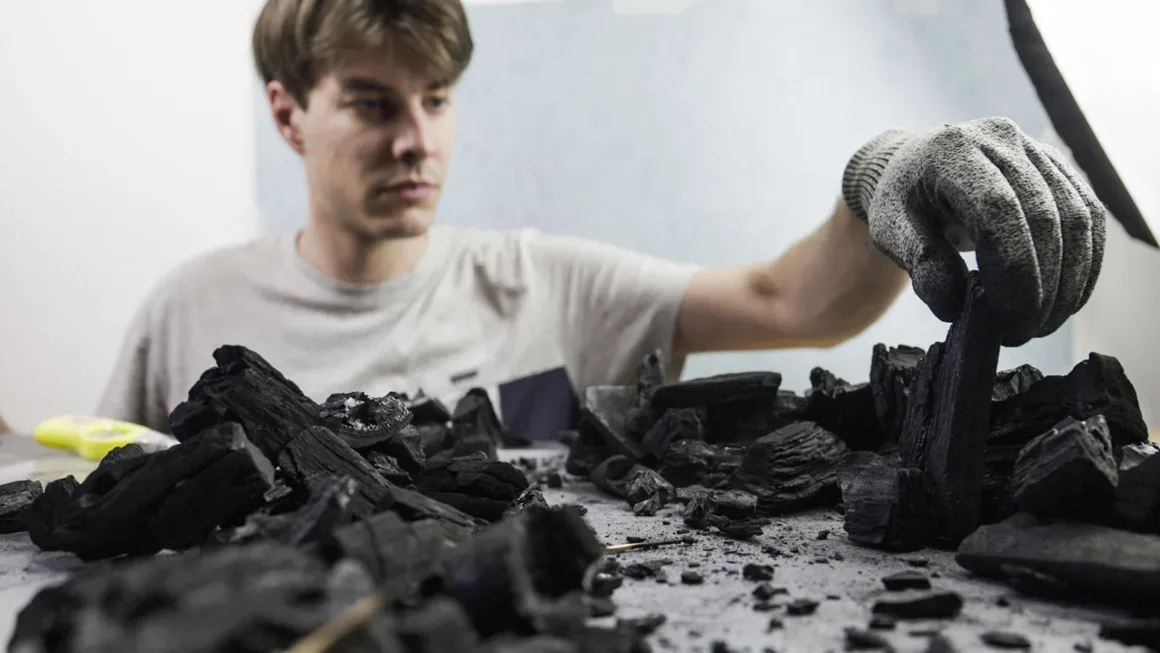 Doğanın Yerini Atıkların Aldığı Bir Dünya: Benedek Lampert'in Yeni Fotoğraf Serisi 11 A person wearing a gray work glove reaches out to place a piece of charcoal onto a pile of charcoal fragments on a table.