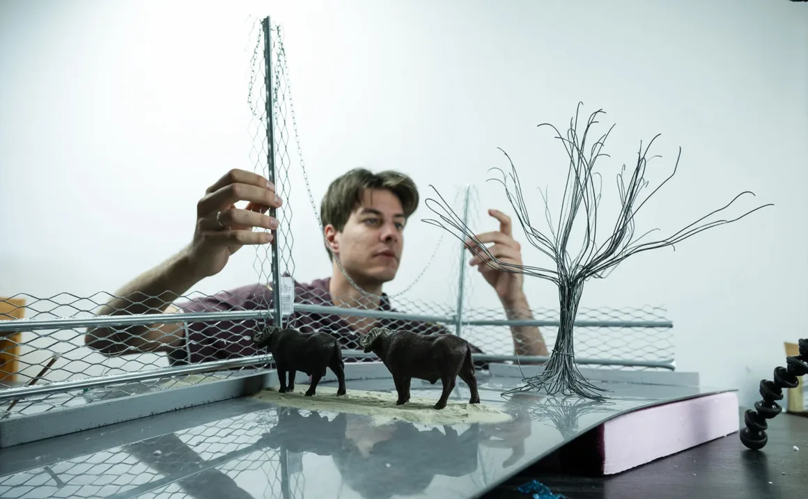 Doğanın Yerini Atıkların Aldığı Bir Dünya: Benedek Lampert'in Yeni Fotoğraf Serisi 15 A person adjusts a miniature wire fence and a wire tree model on a platform featuring two small buffalo figurines.