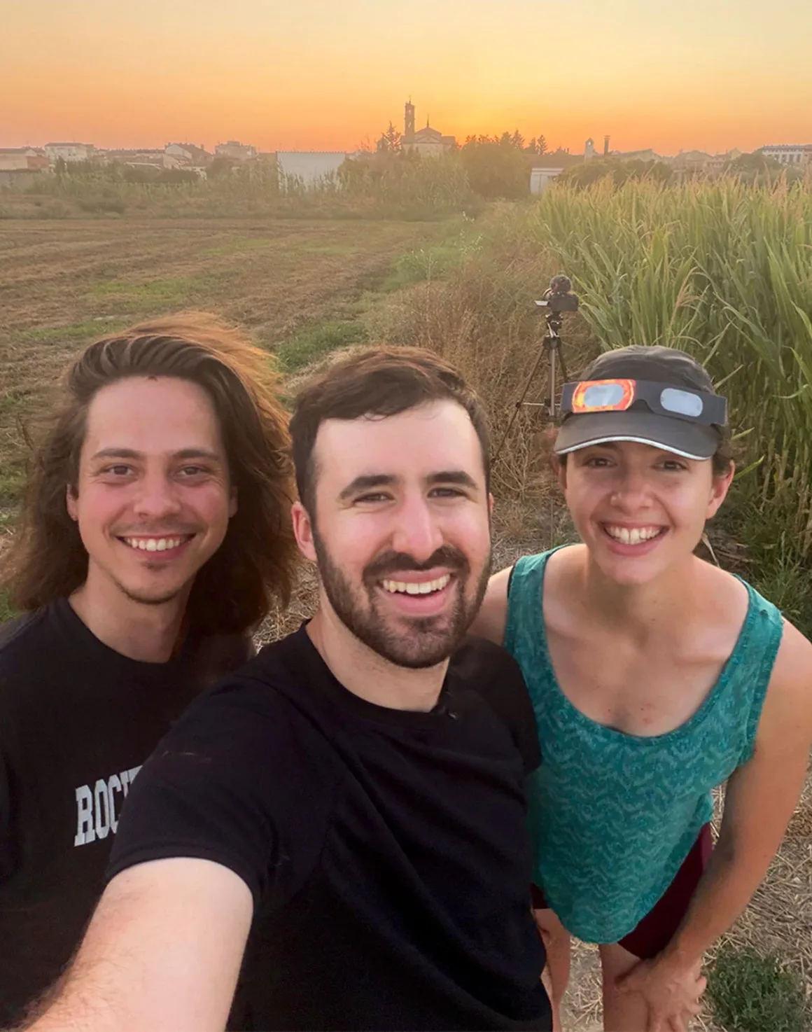 İki Fotoğrafçı Kusursuz Tutulma Karesi İçin Bir Yıl Hazırlandı, Her Şey 30 Saniyeye Kaldı 32 Üç kişi gün batımında bir tarlada selfie için gülümsüyor; arka planda tripod üzerindeki bir fotoğraf makinesi görülüyor.