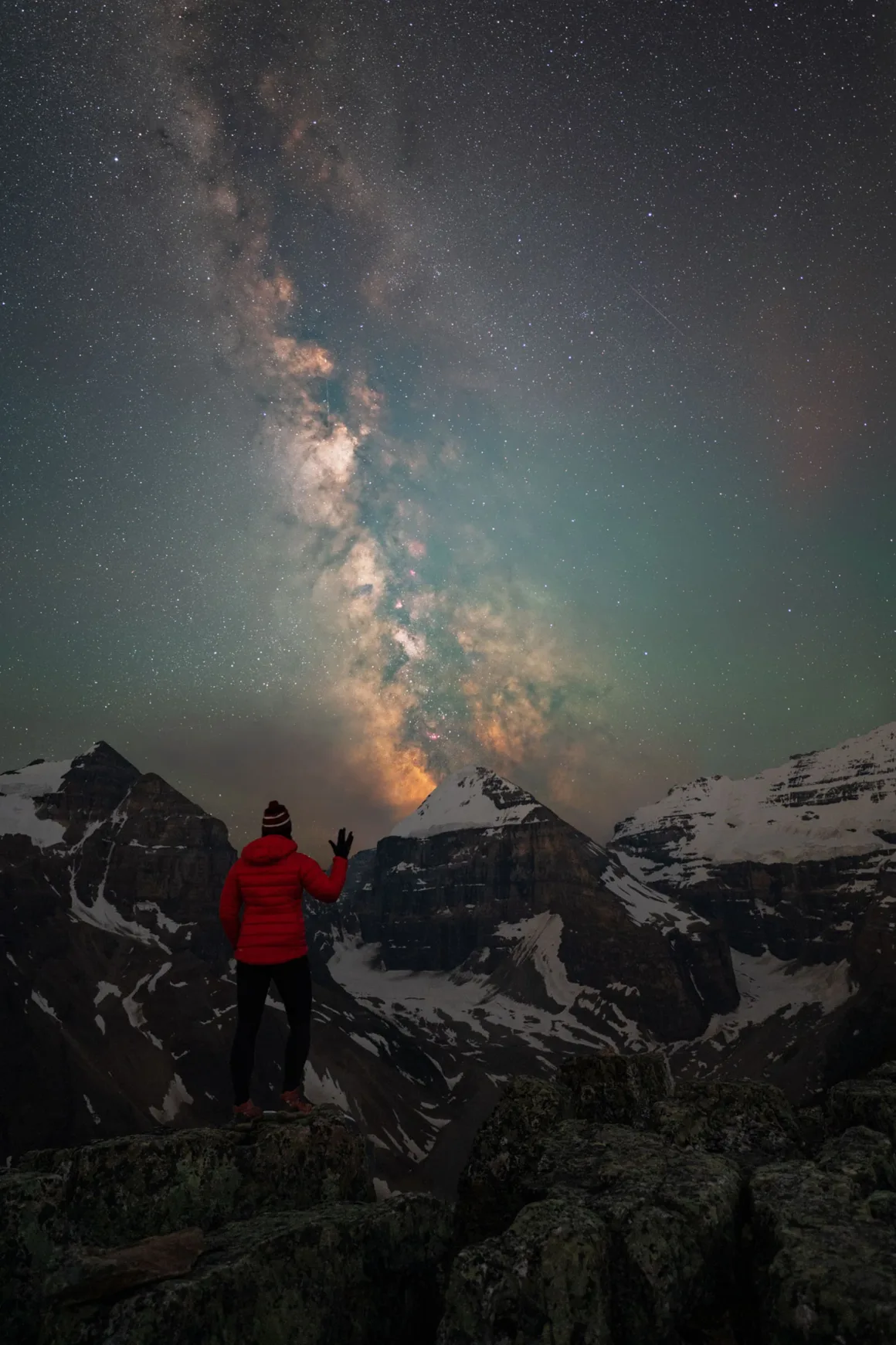 Dünyanın En Karanlık Gökyüzlerinin Güzelliğini Yansıtan 31 Ödüllü Fotoğraf 31 A person in a red jacket stands on a rocky mountain peak at night, gesturing toward the bright Milky Way galaxy visible in the starry sky above snow-capped mountains.