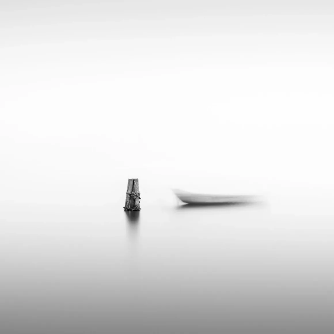 Minimalist Fotoğraf Ödülleri 2026: Kazananlar Sadeliğin Gücünü Gösteriyor 41 A weathered wooden post stands in calm water next to a blurred, out-of-focus boat in a minimalist black and white scene.