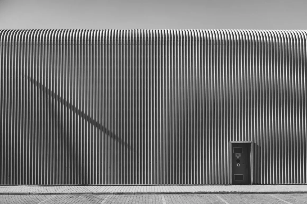 Minimalist Fotoğraf Ödülleri 2026: Kazananlar Sadeliğin Gücünü Gösteriyor 48 A black and white view of a building with a corrugated metal facade, a curved roofline, and a single door on the right side.