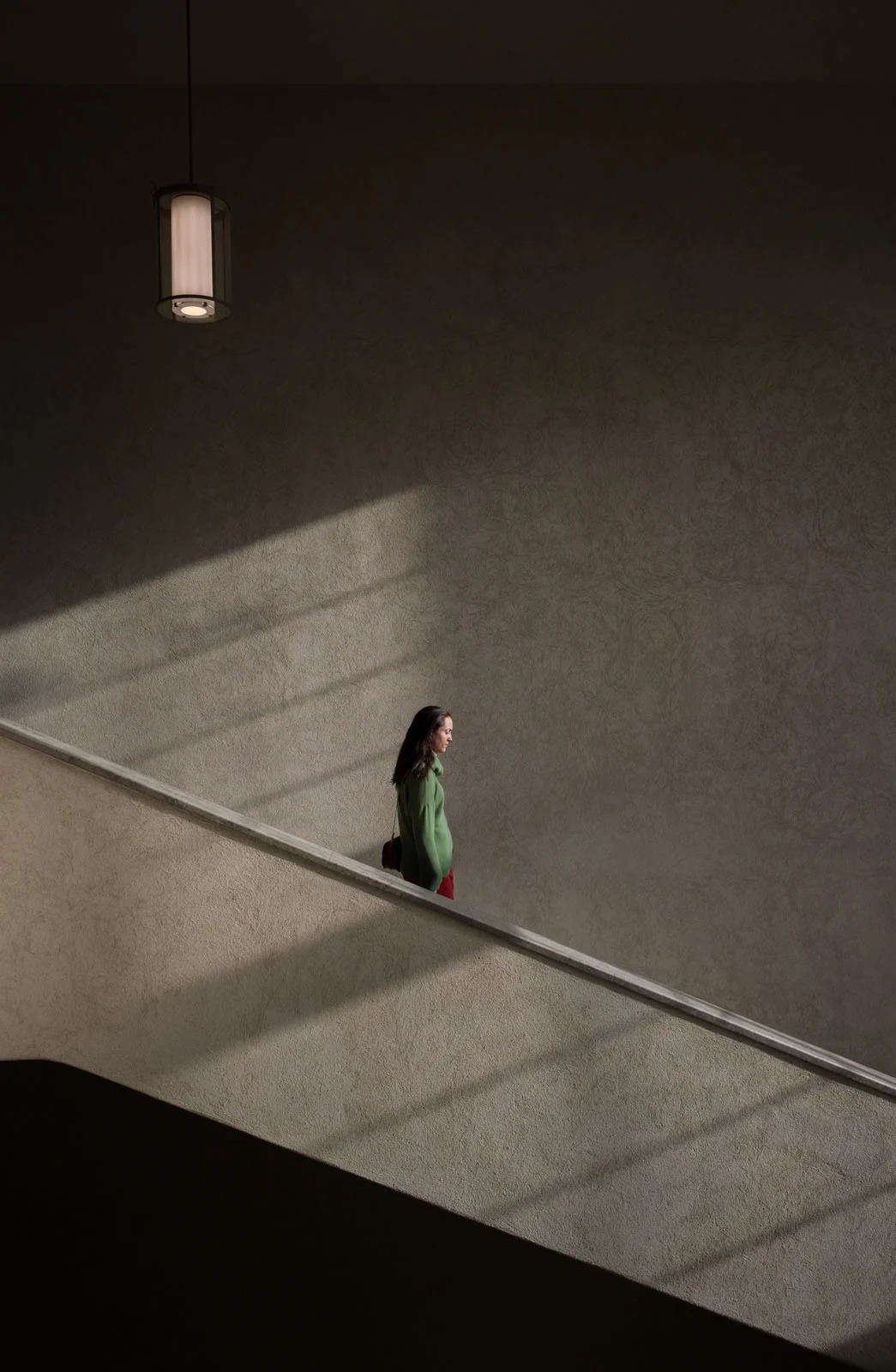 Minimalist Fotoğraf Ödülleri 2026: Kazananlar Sadeliğin Gücünü Gösteriyor 53 A woman in a green sweater walks down a staircase against a textured wall illuminated by a hanging light fixture and diagonal shafts of light.