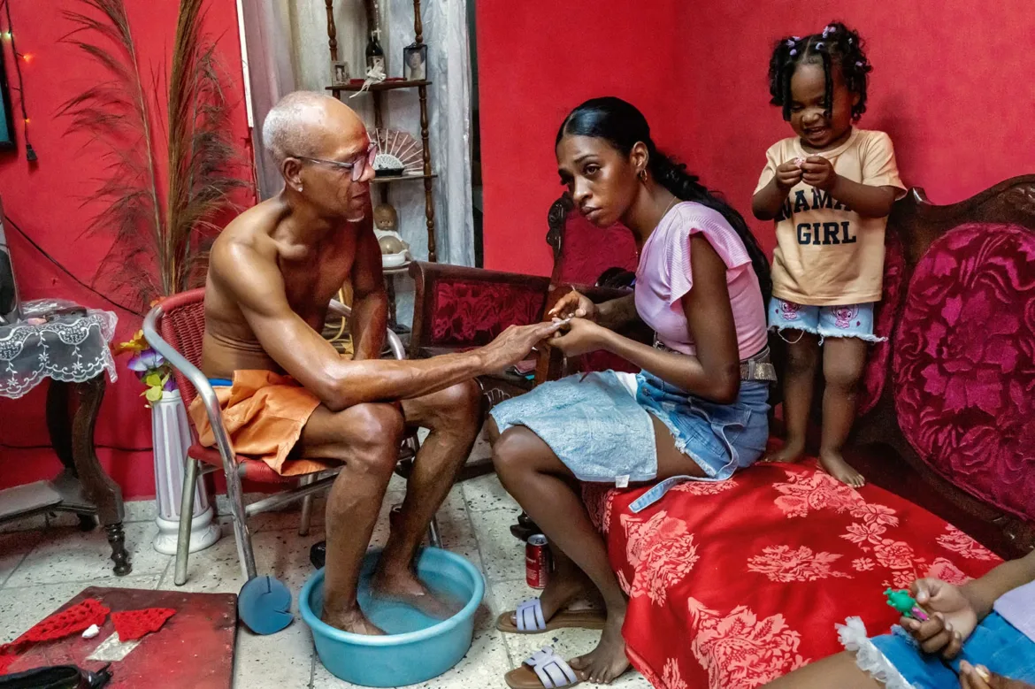 A young woman gives a manicure to an older man who has his feet soaking in a blue basin, while a small child stands nearby.