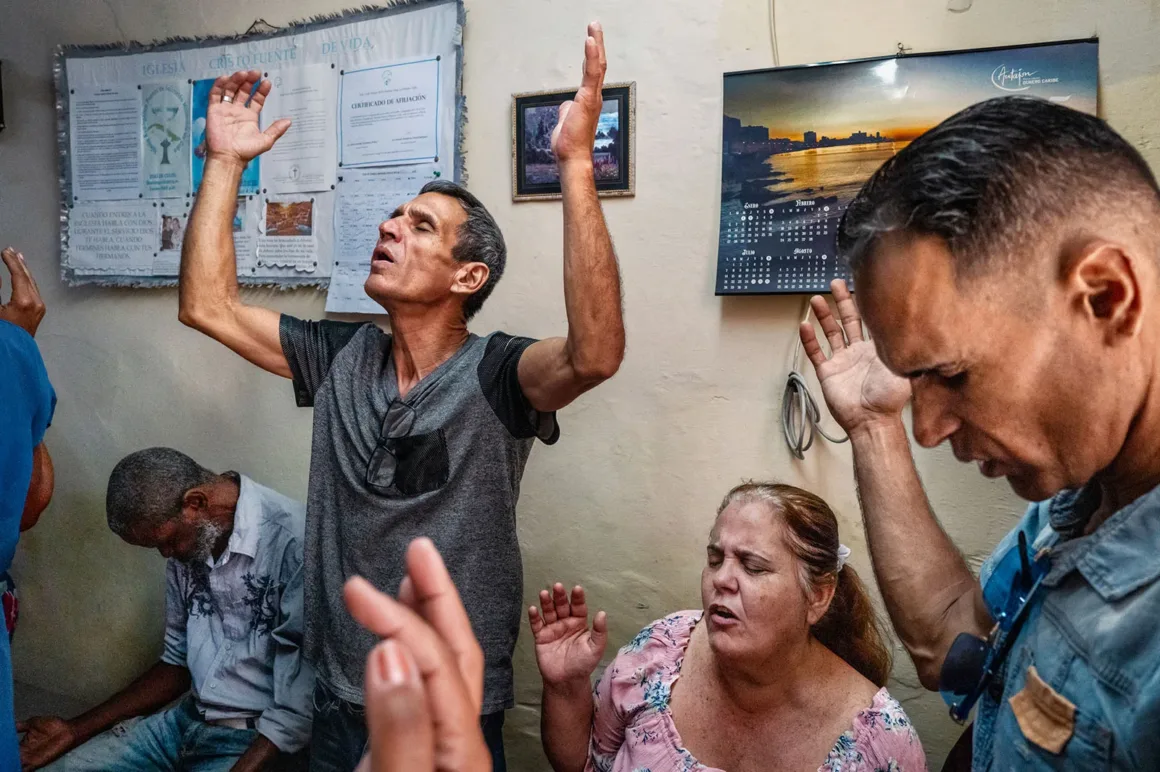Küba’da Dışarıda Kaos, Evlerin İçinde Şefkat 24 Bir grup insan kapalı bir mekânda ellerini kaldırarak dua ederken, yakındaki bir adam başını öne eğmiş halde oturuyor.