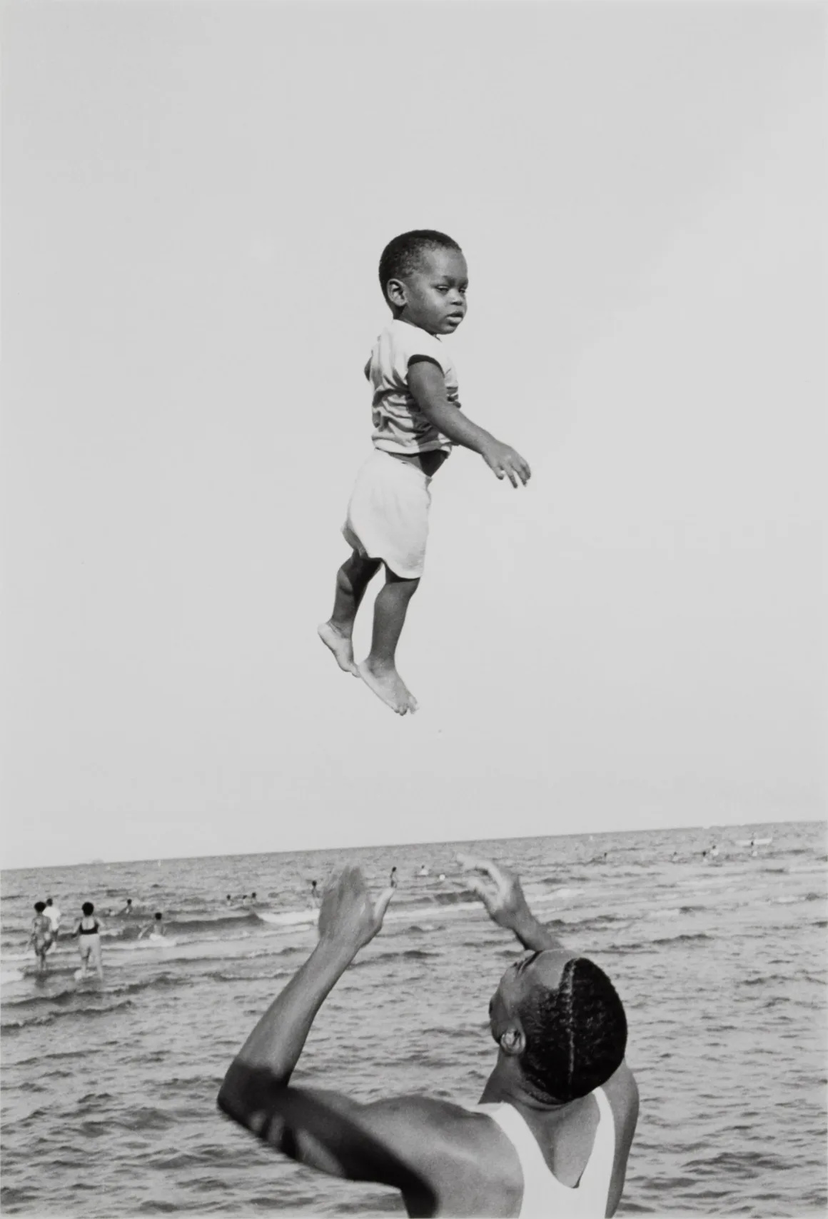 Fotoğraf Sergisi Emmett Till’in Yaşayabileceği Hayatı Düşlüyor 20 Beyaz atletli bir adam küçük bir çocuğu denizin üzerinde havaya kaldırıyor; arka planda insanlar yüzüyor.