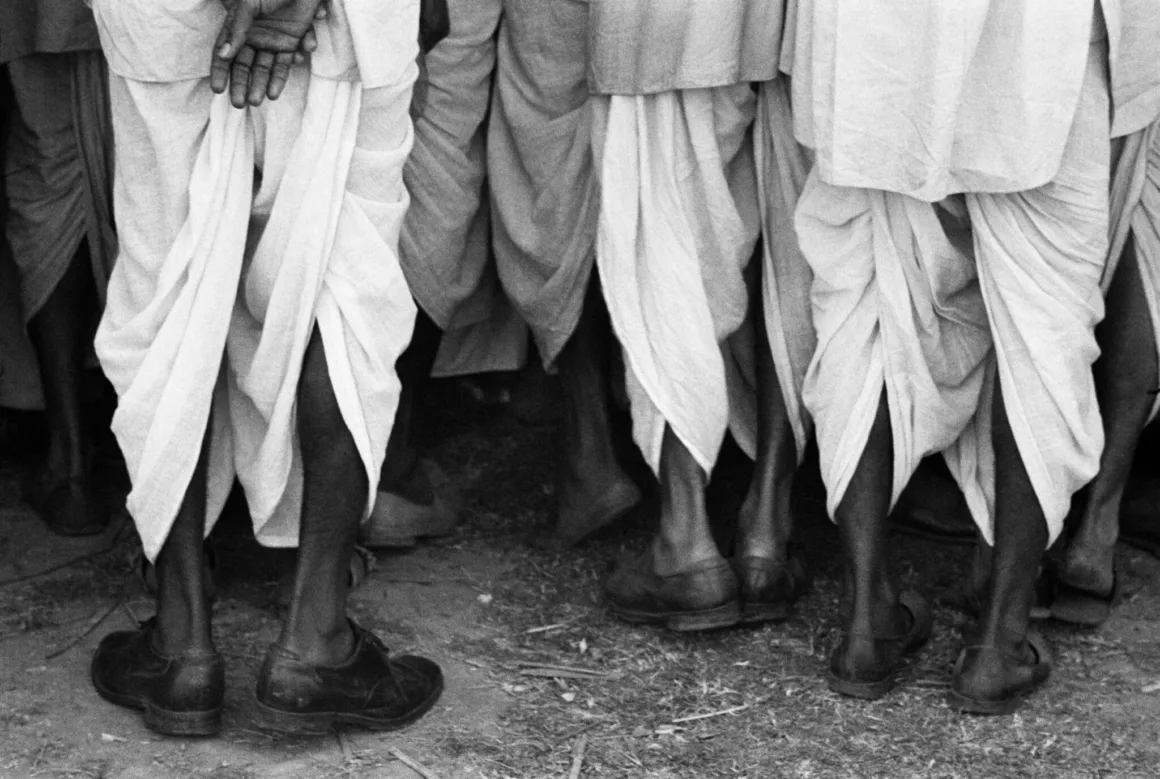 Alçak açıdan çekilmiş siyah-beyaz karede geleneksel dhoti ve koyu deri ayakkabı giyen birkaç erkeğin bacakları görülüyor.