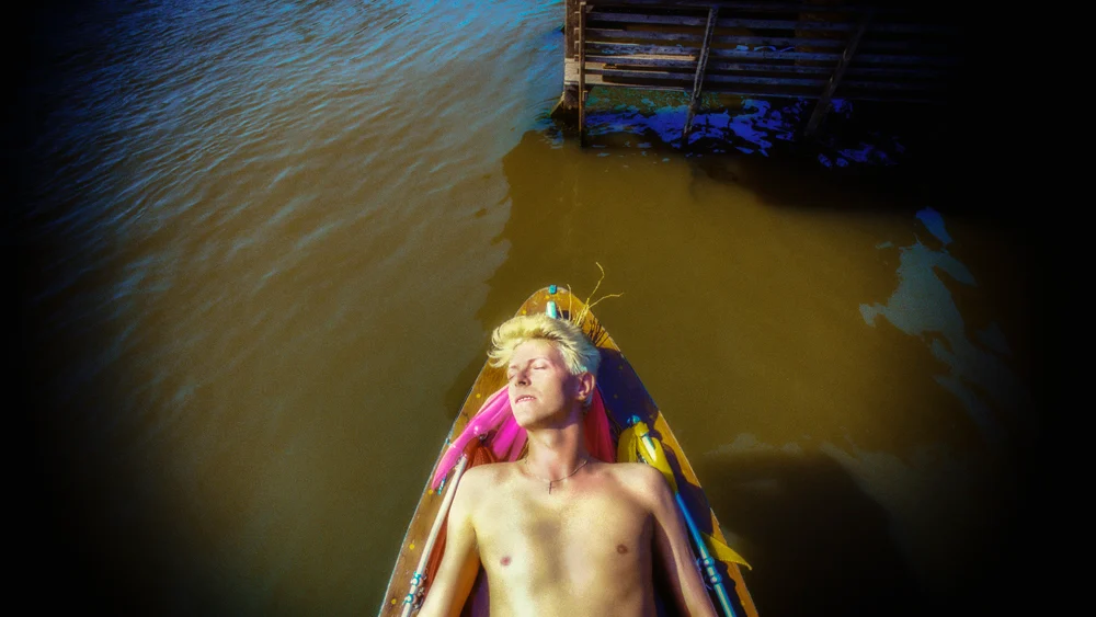 David Bowie Fotoğrafları Sürükleyici Bir Deneyime Dönüşüyor 18 Sarı saçlı David Bowie, Bangkok’ta bulanık su üzerindeki küçük ahşap teknede gözleri kapalı şekilde uzanıyor.