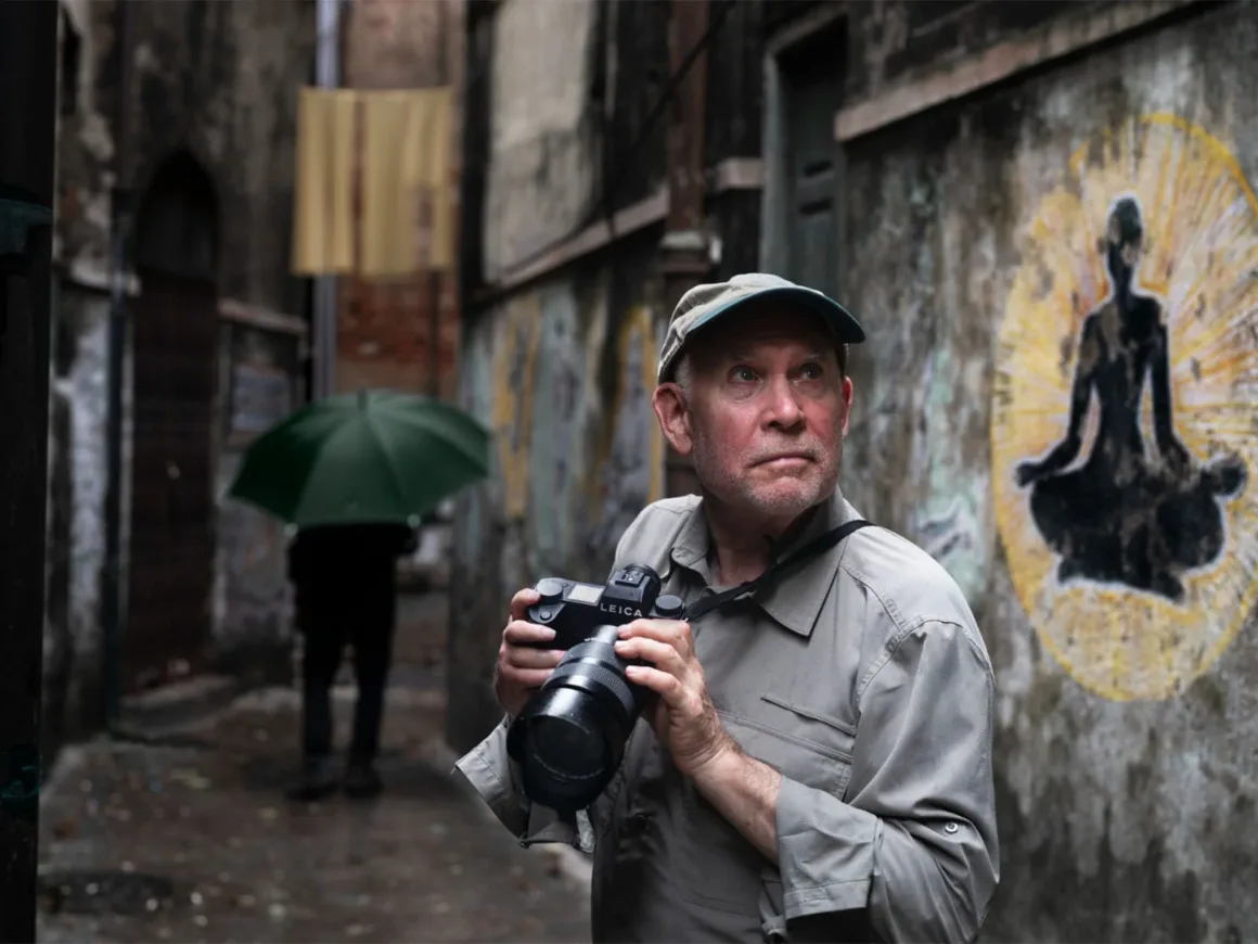 Leica Lux 2.0’a Steve McCurry İmzalı Yeni Görünüm Geldi 19 Şapkalı ve açık renk gömlekli bir adam, oturan bir figürün duvar resminin bulunduğu dar bir sokakta fotoğraf makinesi tutuyor.