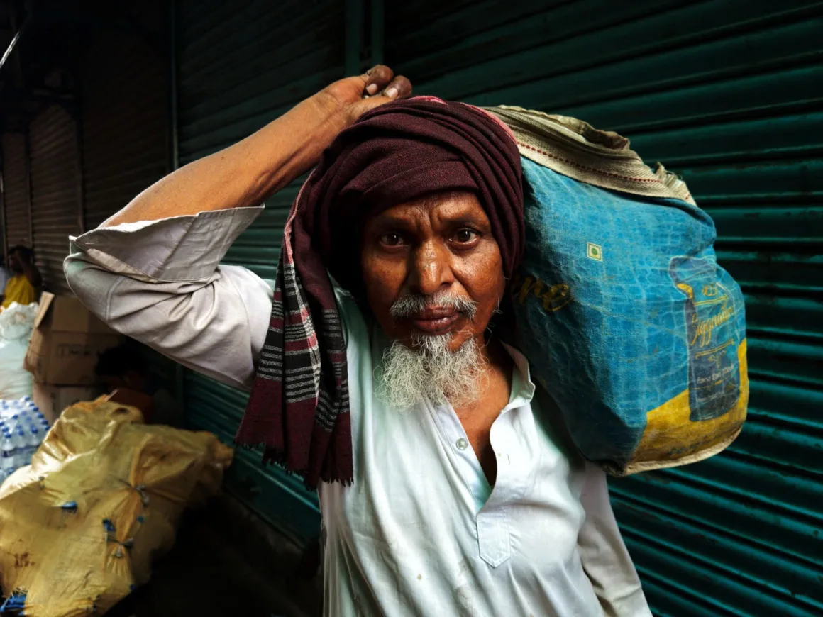 Leica Lux 2.0’a Steve McCurry İmzalı Yeni Görünüm Geldi 21 An elderly man with a white beard and a maroon head wrap carries a large blue and yellow sack over his shoulder while standing in front of a green metal shutter.