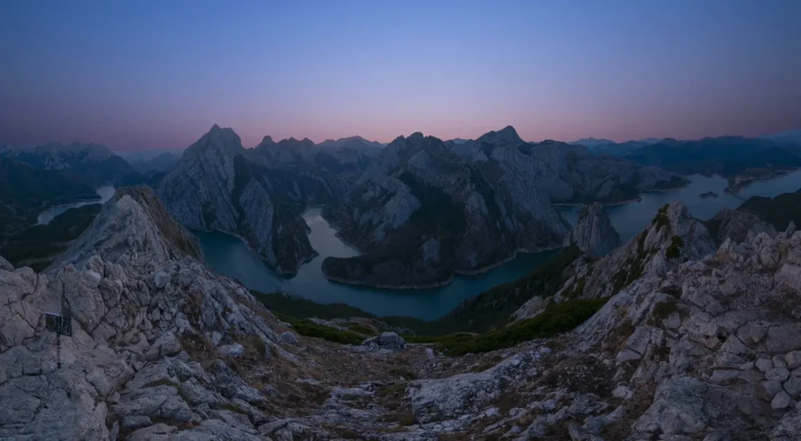 Angel Fux, Tam Güneş Tutulmasını Tek Bir Fotoğrafta Zamana Dönüştürdü 22 A panoramic view of a winding river reservoir surrounded by jagged, rocky mountain peaks under a twilight sky.