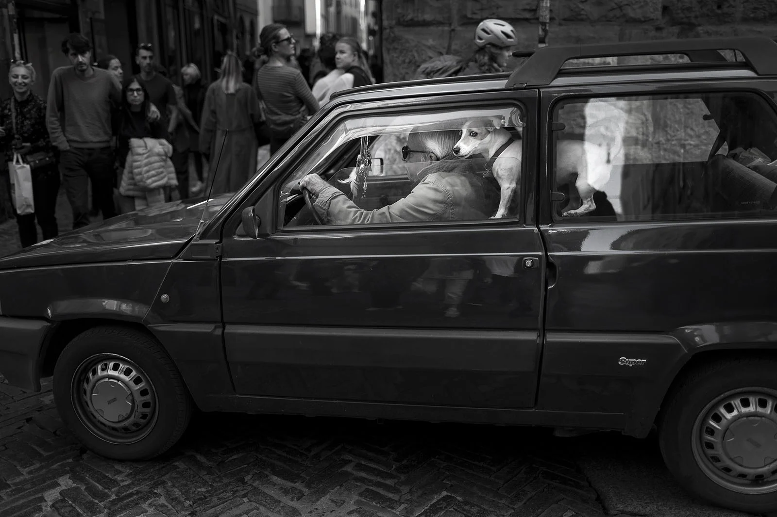 Sony World Photography Awards 2027’ye Katılmanız İçin 15 İlham Verici Fotoğraf 33 A person with a small dog in the passenger seat drives a dark-colored Fiat Panda down a cobblestone street lined with pedestrians.