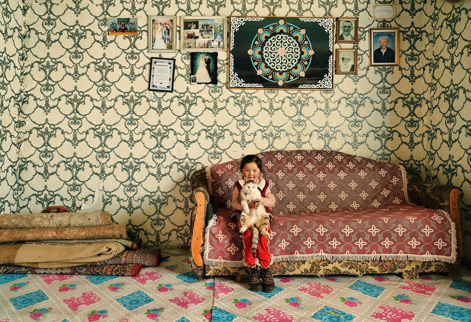 Sony World Photography Awards 2027’ye Katılmanız İçin 15 İlham Verici Fotoğraf 32 A young girl sits on a patterned sofa holding a cat, with a stack of rolled rugs to her left and several framed photos and a decorative wall hanging displayed on the wallpapered wall behind her.