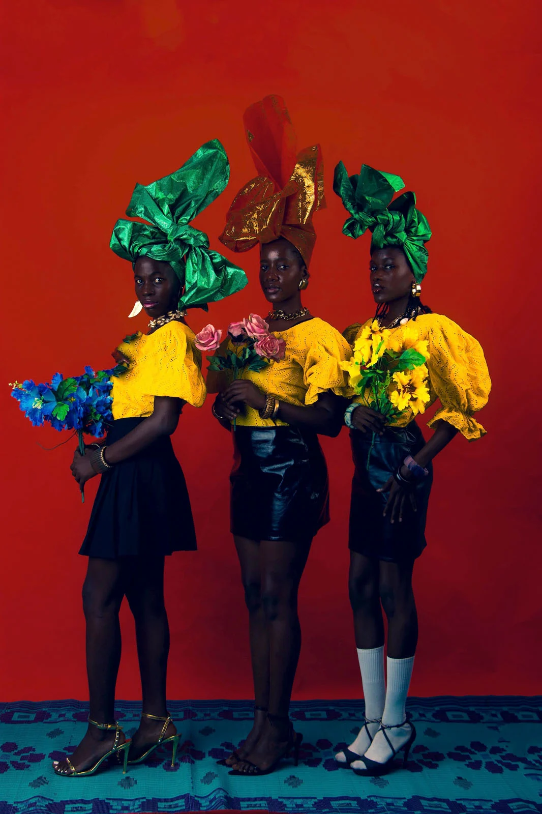 Sony World Photography Awards 2027’ye Katılmanız İçin 15 İlham Verici Fotoğraf 29 Three women stand in a row against a red background, wearing yellow tops, black skirts, and colorful headwraps while holding bouquets of flowers.