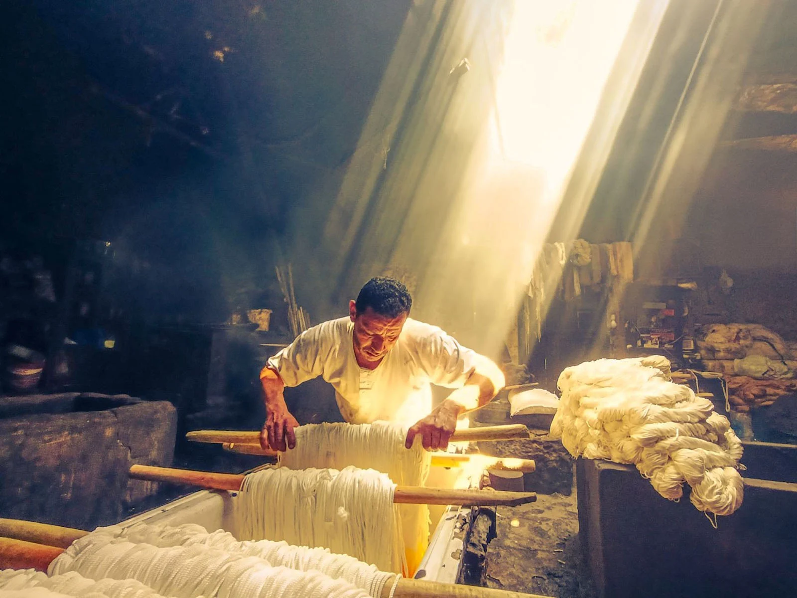 Sony World Photography Awards 2027’ye Katılmanız İçin 15 İlham Verici Fotoğraf 21 A man in a white shirt works with bundles of white yarn over vats in a dimly lit room illuminated by a bright shaft of light.